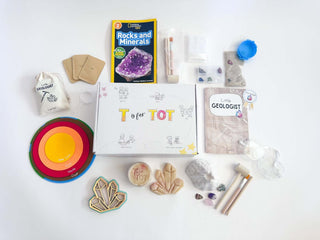 Geologist Kit flat lay including all of the items inside this kit (book, volcano, digging items, gem and playdough, goggles, and felt mat of earth